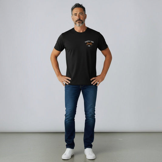 Man wearing the Cigar & Ash Co. Classic Tee in black with crossed-cigars chest logo, blue jeans, and white sneakers against a studio backdrop.