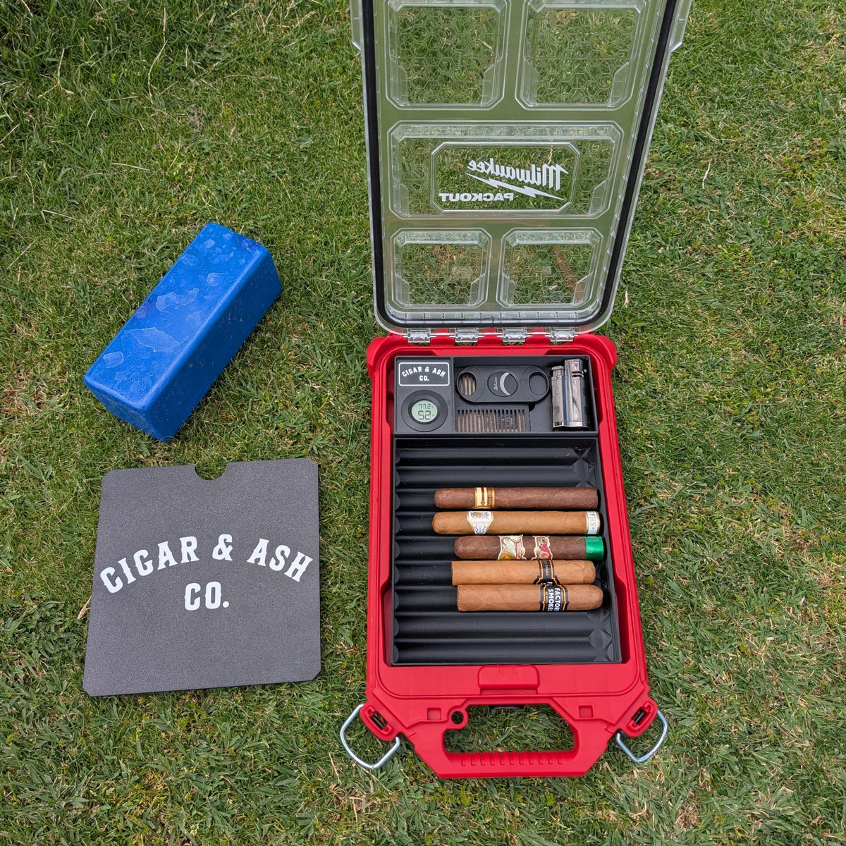 Cigar & Ash Co. Packidor travel humidor kit filled with premium cigars inside a red Milwaukee Packout organizer case set on a golf tee box grass