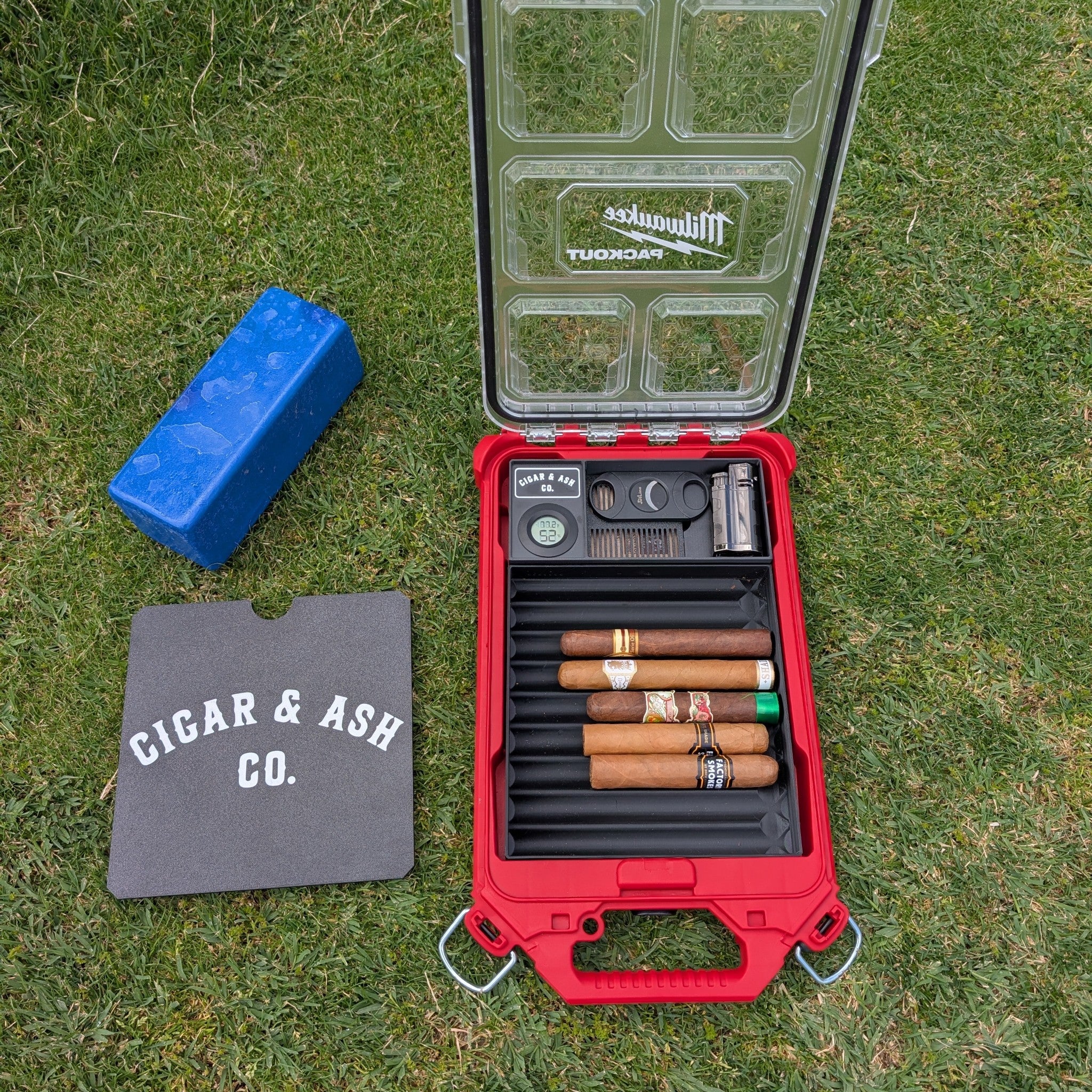Cigar & Ash Co. Packidor travel humidor kit filled with premium cigars inside a red Milwaukee Packout organizer case set on a golf tee box grass