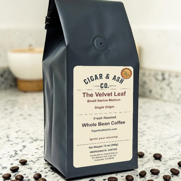 Cigar & Ash Co. The Velvet Leaf Brazil Santos Medium Single Origin whole bean coffee bag sitting on a granite kitchen countertop surrounded by scattered roasted coffee beans.