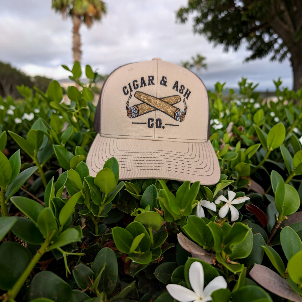 Cigar & Ash Co. beige and brown trucker hat with embroidered crossed cigars logo, set in lush green foliage.
