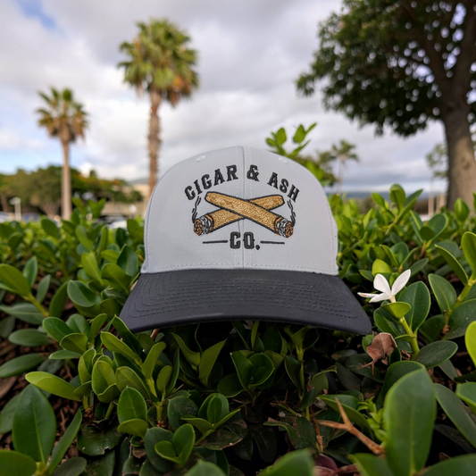Cigar & Ash Co. gray and black laser perforated snapback hat with embroidered cigar logo in a garden setting.