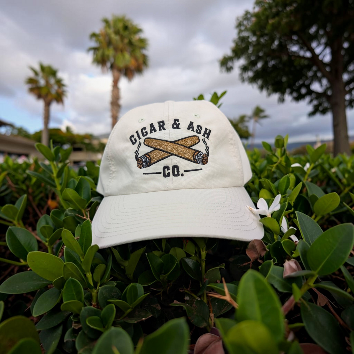Cigar & Ash Co. white relaxed performance hat with embroidered cigar logo resting in green garden foliage.