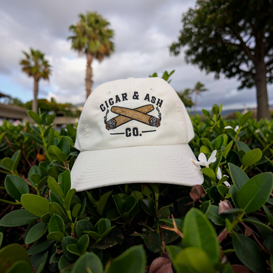 Cigar & Ash Co. white relaxed performance hat with embroidered cigar logo resting in green garden foliage.